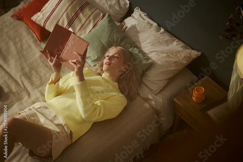 Foto Young Caucasian woman lying on bed and reading book, relaxing at home