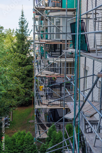 Long residential building facade covered with scaffolding during extensive exterior renovation, with green trees beside the construction site. Concept of housing renewal, property maintenance.