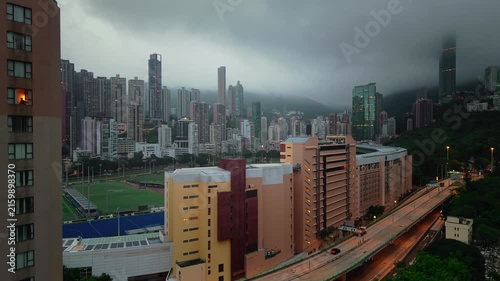 Cinematic aerial view of a Hong Kong residential neighborhood surrounded by skyscrapers and green hills during overcast weather, with city traffic and illuminated buildings.