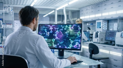 Male scientist in white lab coat sitting at a desk, reviewing complex molecular structures on a curved computer monitor in a bright laboratory