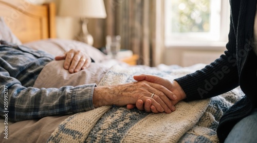 Close-up of a person holding the wrinkled hand of an elderly patient resting in a wooden bed, wearing a plaid shirt and covered by a woven blanket