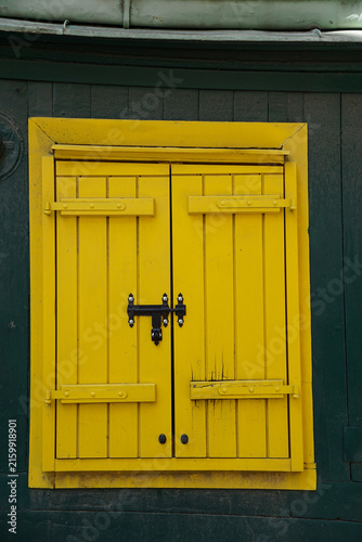 Front view of vibrant yellow wooden shutters with black metal hardware set against a dark green wall, creating bold color contrast and traditional architectural character.