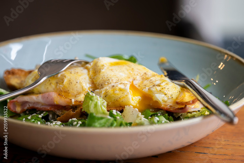 Eggs Benedict with perfectly poached eggs and a runny golden yolk, crispy bacon and creamy hollandaise sauce on toasted bread, served with a fresh green salad and sesame seeds. 