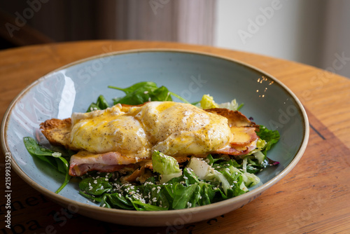 Classic Eggs Benedict served on toasted bread with crispy bacon, poached eggs and creamy hollandaise sauce, accompanied by a fresh green salad sprinkled with sesame seeds.