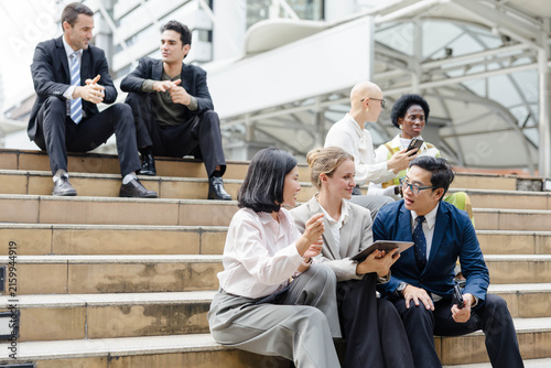 Professionals in business attire networking on outdoor steps, discussing work with a tablet and coffee in a modern urban setting.