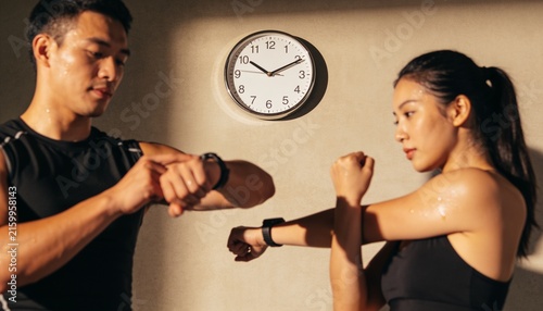 Checking man left, woman right wearing black tops, checking wrist trackers at fitness studio, clock