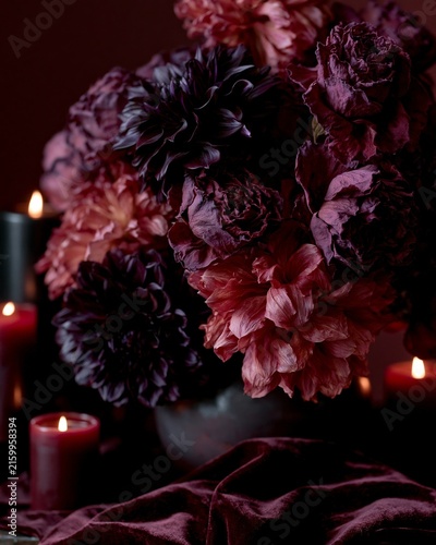 Moody autumn still life featuring a large arrangement of deep purple and pink flowers with candles and rich fabric in a dark setting