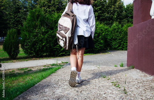 Schoolgirl walks on a pathway leading to school. She carries a backpack and wears a school uniform. It is daytime