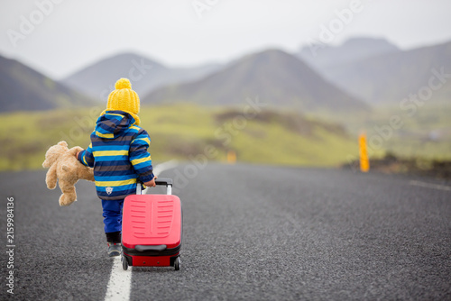 Cute toddler child, boy with teddy bear and suitcase in hand, running on a road in Iceland on a rainy day