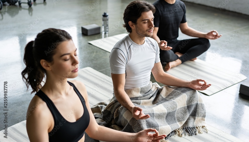 Fototapeta premium Meditating trio in sportswear on striped mats in studio with plaid blanket, water bottle and blocks