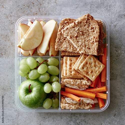School lunch box healthy lunch container with sliced apple, grape, sandwich, cracker, carrot, balanced snack packed in clear container on gray stone table
