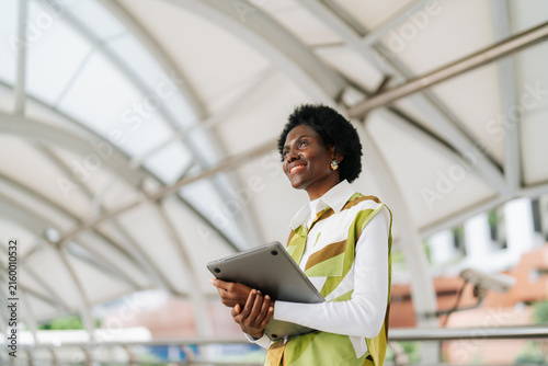 Smiling businesswoman holding laptop looking up hopefully in the city with modern skyscrapers, inspiration, ambition, urban lifestyle and professional confidence concept.