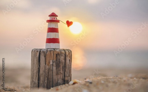 Fotografie Miniature red and white wooden lighthouse toy perched upon a weathered wooden piling with a small dangling heart charm during a beautiful soft golden hour sunset at the beach side