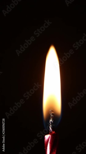 A tall bright candle flame burns steadily against a dark background near a red wick, flickering softly while a sudden spark flares upward before the flame settles calmly again.