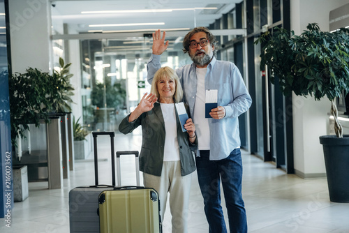 Foto Senior couple with luggage waving and smiling in modern airport terminal, holdin