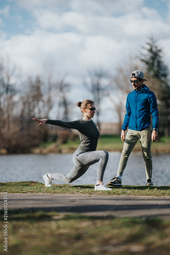 Obraz premium A girl performs a deep lunge while another girl acting as a trainer watches and guides. The outdoor workout takes place on grass near a lake in a park setting.