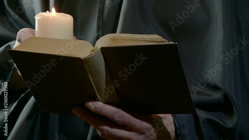 Monk holding candle and reading open book in dim dramatic light