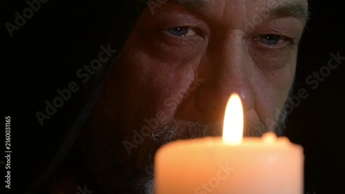 Closeup of hooded monk slowly opening eyes before glowing candle flame