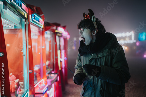Obraz na płótnie Young man wearing a reindeer headband reacts with surprise while playing a claw machine