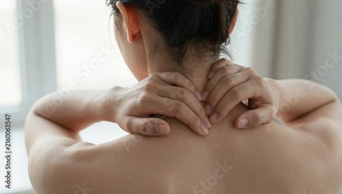 Young woman holding her neck in pain and discomfort at home, grimacing from neck ache and tension while seeking relief and self-care in a bright modern living space