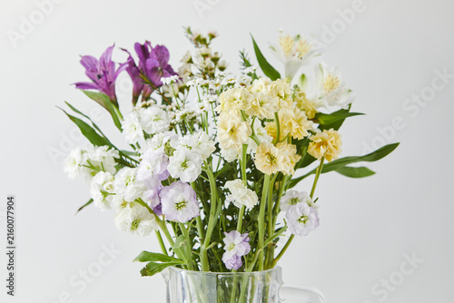 Fresh mixed bouquet of purple alstroemeria and white yellow stock flowers in a clear glass jug. Spring floral arrangement on a light background.