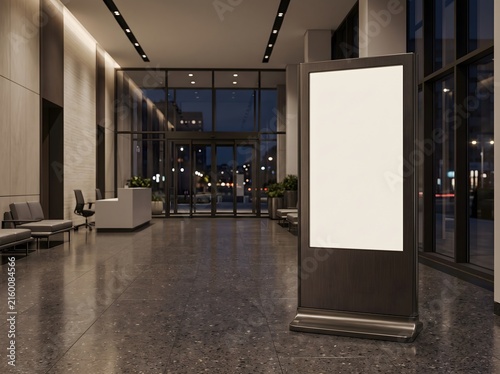 Blank vertical digital signage display standing in a modern office building lobby at night.