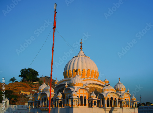 View of the white and yellow domes of Gurudwara Sri Panja Sahib with a tall orange flagpole against a clear blue sky in Hassan Abdal, Punjab, Pakistan.