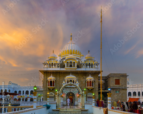View of Gurudwara Sri Panja Sahib with its ornate white and gold dome, intricate architectural details, and a person entering the entrance, set against a sunset sky in Hassan Abdal, Punjab, Pakistan.