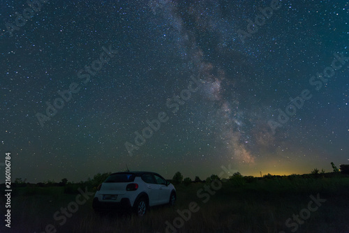 A white car stands in a dark, open field under a spectacular night sky filled with the bright Milky Way