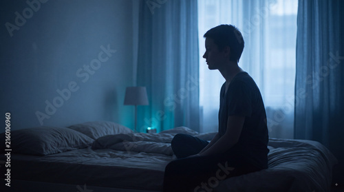 Lonely teenager sitting on bed in dark blue bedroom at dawn, contemplative silhouette by window expressing sadness, insomnia and mental health struggle