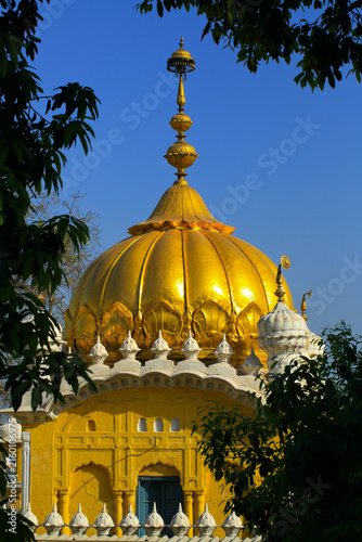 View of the golden dome of Gurdwara Dera Sahib framed by tree leaves against a clear blue sky in Lahore, Punjab, Pakistan.
