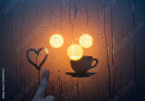 Finger drawing heart and coffee cup silhouette on rainy window pane. Cozy mood with warm bokeh lights in evening. Romantic atmosphere during autumn rain for leisure and relaxation concept.