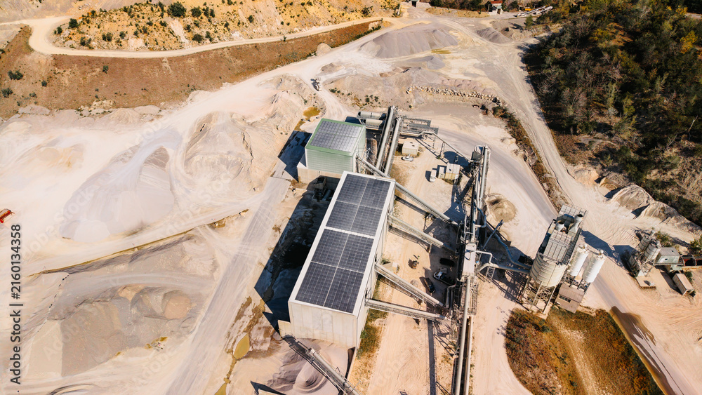 Naklejka premium Aerial view of sand quarry processing facility with solar panels. Modern aggregate plant with rooftop solar panels silos conveyor systems and surrounding terrain.