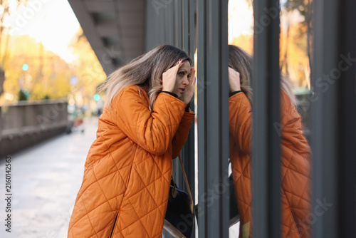 A woman in an orange coat peering through metal bars on an urban sidewalk, evoking a sense of curiosity and movement in the cityscape. The scene captures the essence of urban life with its.