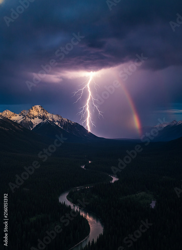 Lightning storm over mountain river landscape. Dramatic rainstorm sky with rainbow over valley. Thunderstorm weather with peak and winding river. Natural phenomenon night sky.