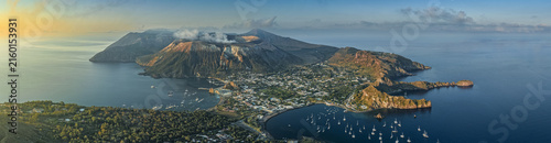Aerial view of the volcanic island of Vulcano with its smoking crater, coastal town, and numerous boats anchored in the bay at golden hour in Lipari, Sicily, Italy.