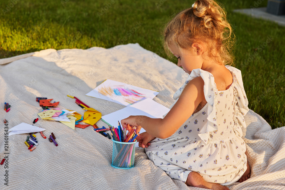 Fototapeta premium A young child sits on a blanket surrounded by crayons and paper. She focuses on her drawing while the sun shines in the background during late afternoon