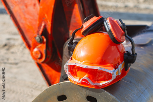 Orange safety helmet with protective goggles and earmuffs placed beside heavy construction equipment, representing workplace safety, industrial work, protective gear and construction industry.
