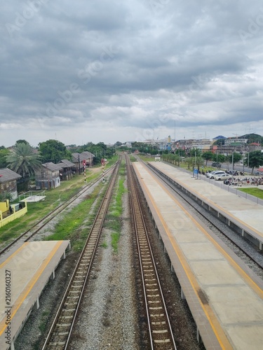 Bild auf Leinwand A high-angle elevated view showing long parallel railway tracks stretching through an empty train station platform with a cloudy sky and rural landscape background