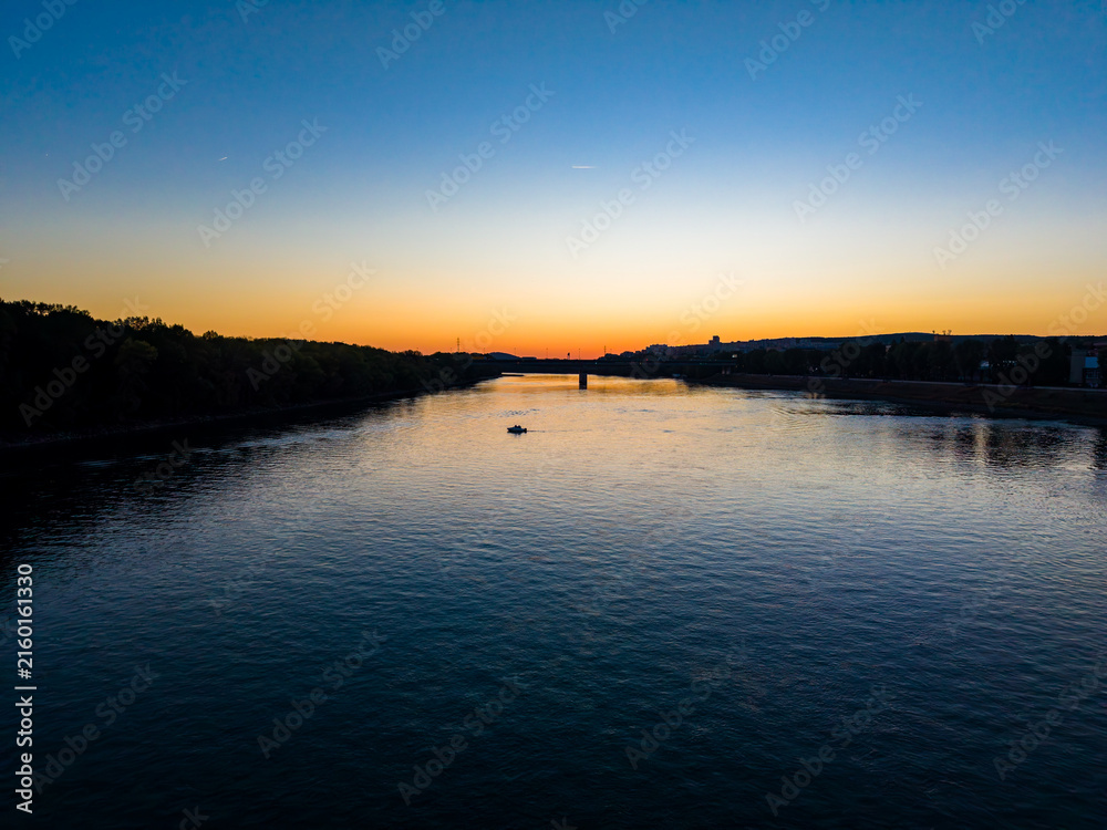 Fototapeta premium River at sunset with a small boat in the water and hills in the background near a city along the riverbank