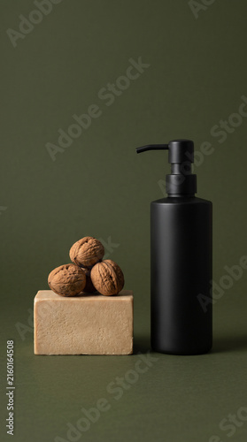 Matte black cosmetic pump bottle mockup next to walnuts on stone pedestal against olive green background