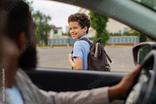 Black schoolboy with backpack looking back at father driving him to school