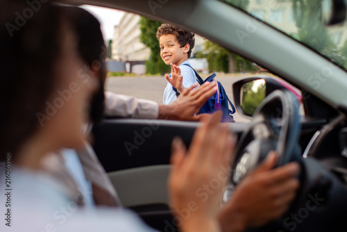 Smiling schoolboy waving goodbye to parents from outside car before heading school