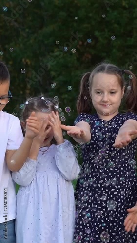 Happy children play with soap bubbles and laugh in summer park. Girls and boys catch bubbles. Team kids play outdoors. Childrens leisure. Vertical shot