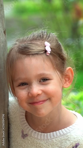 Portrait of little girl looks at camera and laughs on playground. Happy child plays in green summer park. Kid leisure outdoors. Vertical shot