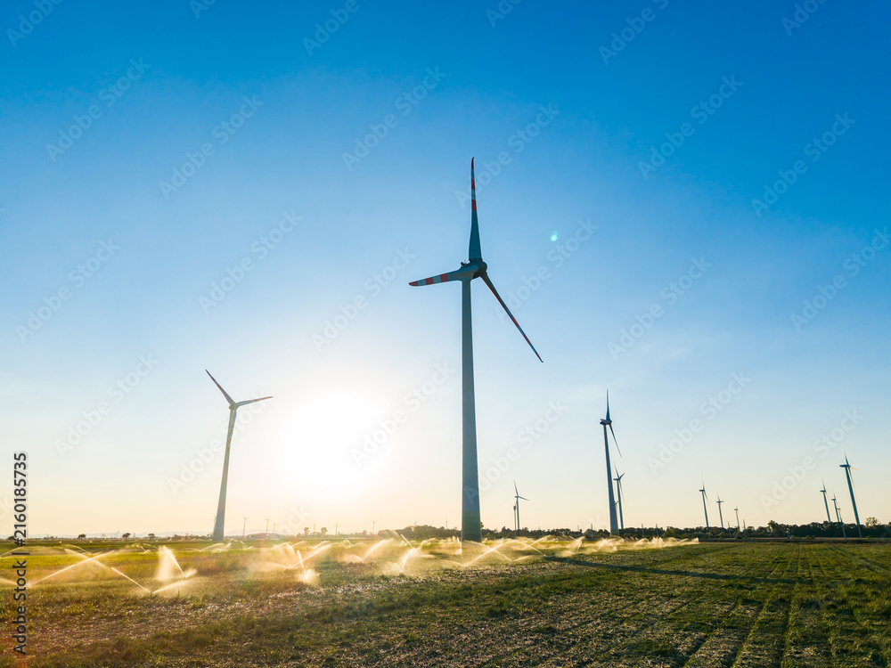 Obraz premium Wind turbines stand tall over fields with sprinklers operating at sunset near a farming area