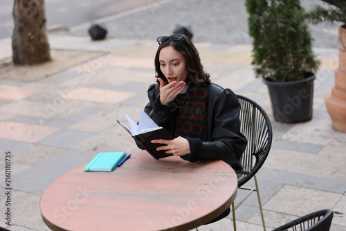 Young woman sitting at outdoor caf table reading open notebook with surprised expression, hand near mouth, pen and second notebook beside her, in soft natural daylight with blurred street background