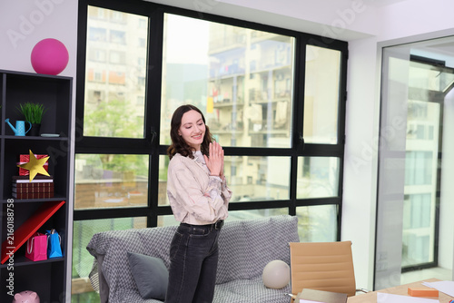 Smiling young woman stands indoors with hands clasped beside sofa and shelving in bright modern apartment, with large windows, soft daylight, colorful decor, and calm relaxed home setting