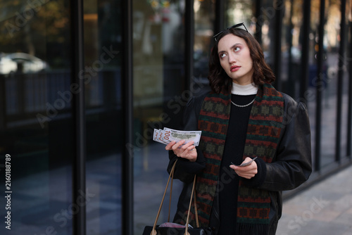 Thoughtful young woman standing beside glass storefront on city sidewalk, holding cash and cards while looking upward, wearing dark outfit with patterned scarf, soft natural light and shallow depth