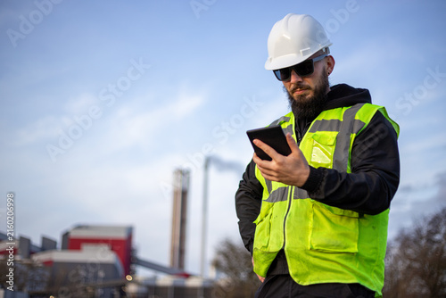 Engineer worker wearing hard hat and safety vest, looking at digital tablet in front of industrial factory